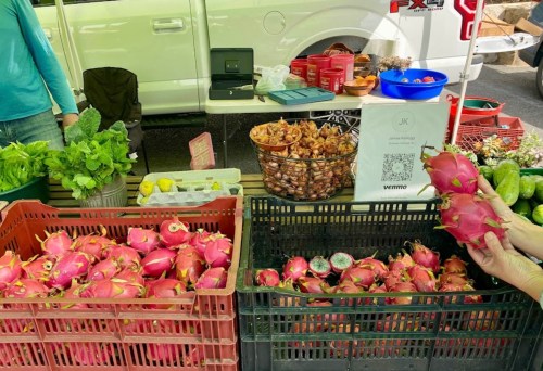 A market stall displaying fresh produce, including dragon fruit, herbs, and various vegetables in crates.