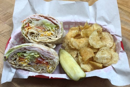 A basket with two wraps filled with turkey, lettuce, and cheese, served with potato chips and a pickle.