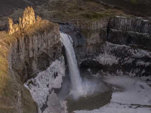 A stunning waterfall cascades down rocky cliffs, surrounded by icy terrain and illuminated by soft sunlight.