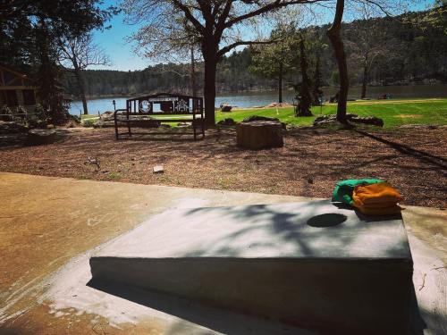 A serene lakeside view with a concrete platform, trees, and a picnic area in the background under a clear blue sky.
