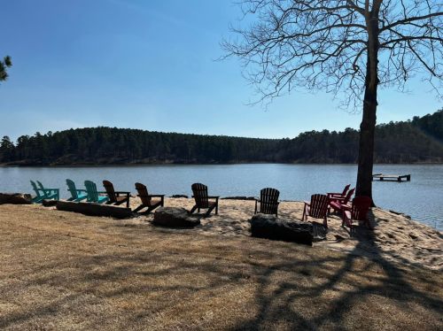 A serene lakeside view with colorful chairs arranged on the shore, surrounded by trees and a clear blue sky.