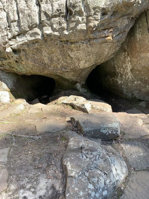 A rocky cave entrance surrounded by large stones and earthy ground, with sunlight illuminating the scene.