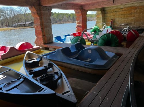 Colorful paddle boats and canoes are docked under a wooden structure by a calm lake.