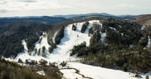 Aerial view of a snowy hillside with ski slopes, surrounded by trees and mountains under a cloudy sky.