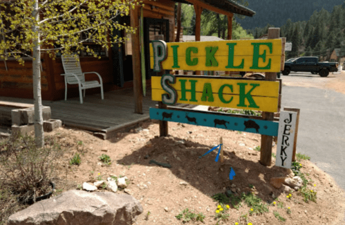 Colorful wooden sign for "Pickle Shack" with a "Jerky" sign below, set in a rustic outdoor location.