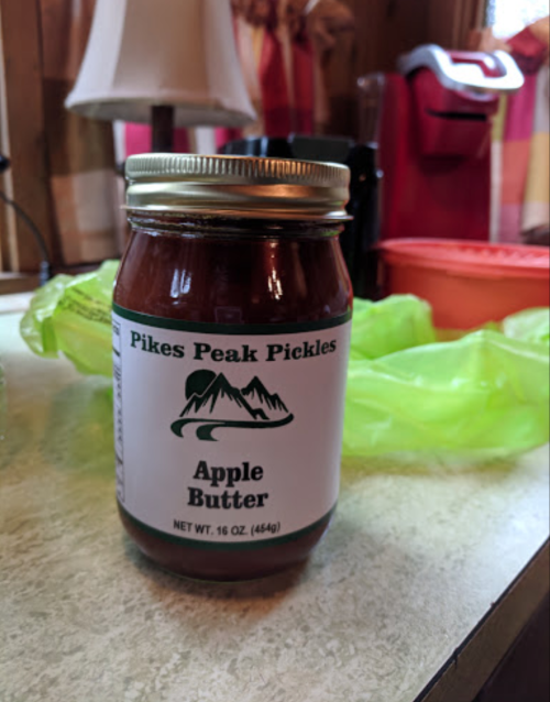 A jar of Pikes Peak Pickles apple butter on a countertop, with a lamp and other kitchen items in the background.