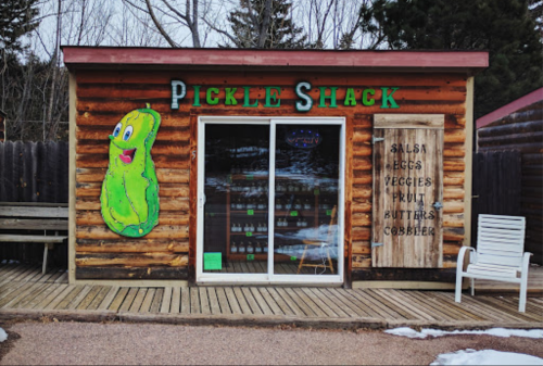 A rustic wooden building with a large pickle mural, labeled "Pickle Shack," featuring signs for salsa, veggies, and fruit.
