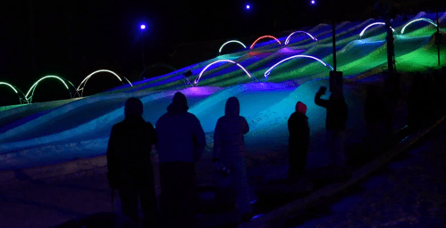 People stand in silhouette against a brightly lit snow tubing hill with colorful lights at night.