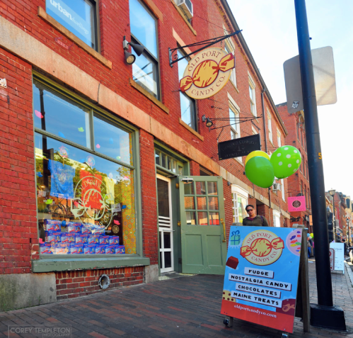 A colorful candy shop with a sign, balloons, and a sidewalk display of treats in a brick building.