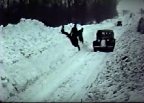A black-and-white scene of a snowy road with a person slipping and falling near a vintage car. Snowbanks line the sides.