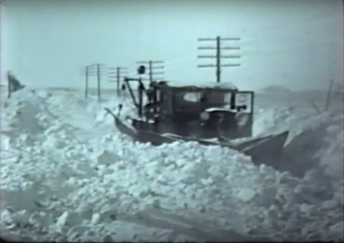 A vintage snowplow clears a snow-covered road, surrounded by tall snowdrifts and telephone poles in the background.