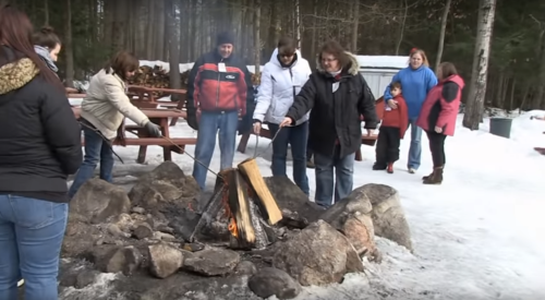 A group of people gathered around a campfire in a snowy forest, roasting food on sticks.
