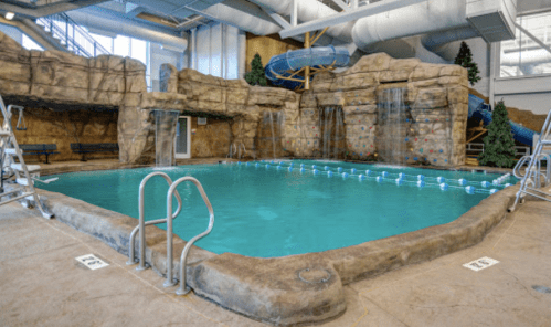 Indoor pool with rock formations, waterfalls, and a water slide, surrounded by benches and trees.