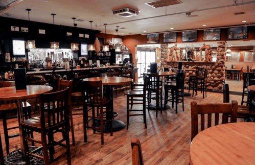 A cozy restaurant interior with wooden tables, chairs, a bar, and stone accents, featuring warm lighting and a welcoming atmosphere.