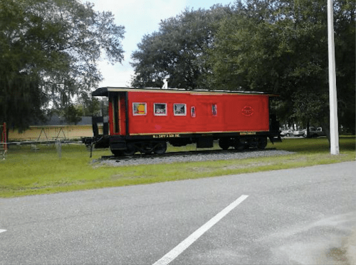 A red caboose on display near a grassy area, with trees and a playground in the background.