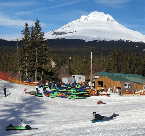 A snowy landscape with people tubing, a mountain in the background, and trees lining the area.