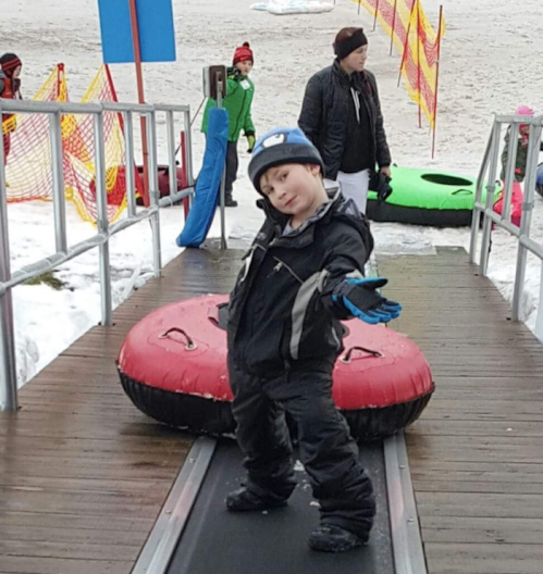 A child in winter gear poses playfully on a snowy ramp, with snow tubes and other people in the background.