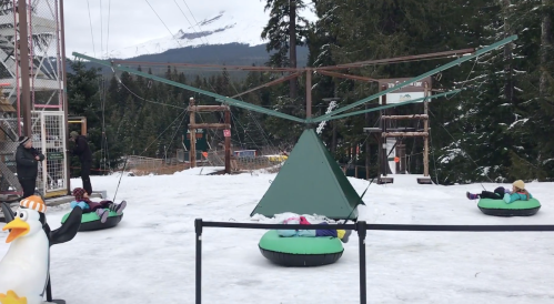 Children enjoy snow tubing on a snowy hill, with a green triangular structure and trees in the background.