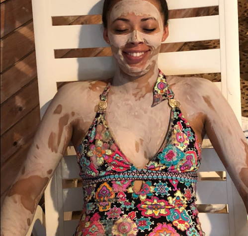 A woman relaxes on a lounge chair with a facial mask, wearing a colorful swimsuit, smiling contentedly.
