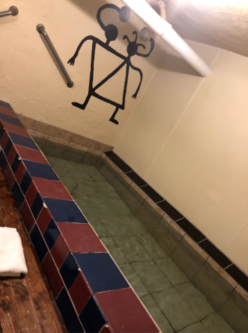 A tiled pool area with a mural of two abstract figures on the wall. A towel is placed on a wooden ledge nearby.