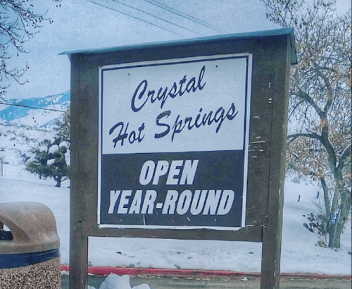 Sign for Crystal Hot Springs, stating "Open Year-Round," with a snowy landscape in the background.