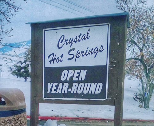 Sign for Crystal Hot Springs, stating "Open Year-Round," with a snowy landscape in the background.