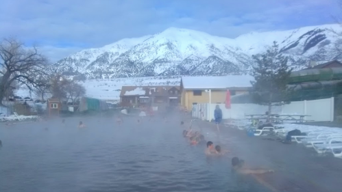 A steaming hot spring surrounded by snow-covered mountains and a few people enjoying the warm water.