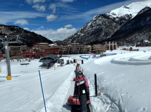 A snowy landscape with people tubing, surrounded by mountains and a ski resort in the background under a blue sky.