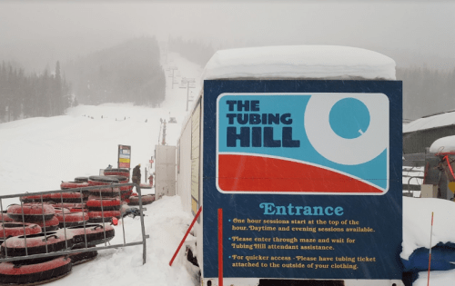Sign for "The Tubing Hill" entrance, with snow-covered hills and tubing equipment in the background.