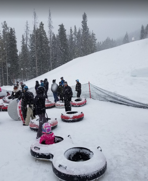 People enjoying a snowy day at a tubing hill, surrounded by snow-covered trees and tubing equipment.