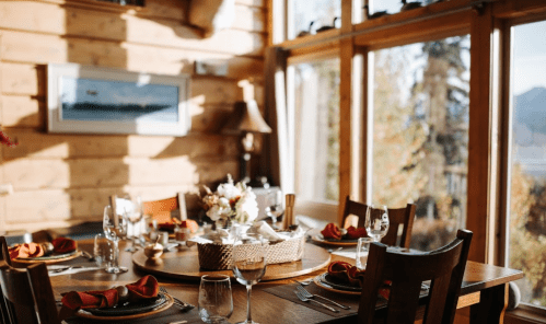 A cozy dining table set with elegant tableware, surrounded by warm wooden walls and large windows showcasing nature.