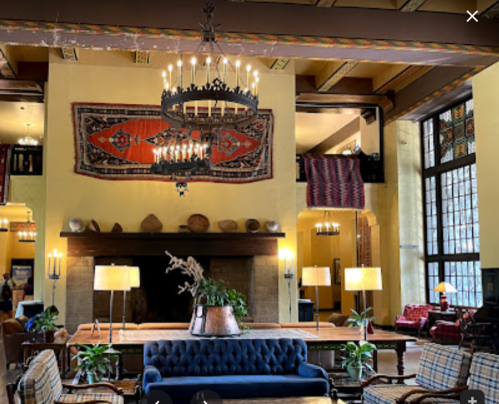 A cozy lobby with a large chandelier, colorful rugs, and comfortable seating, featuring warm lighting and plants.