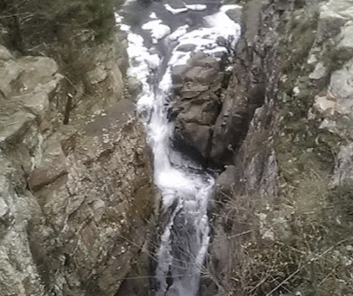 A narrow canyon with a waterfall cascading between rocky cliffs, surrounded by sparse vegetation.