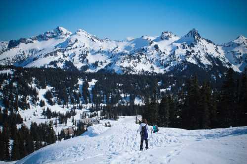 A person hiking on a snowy trail with mountains in the background under a clear blue sky.