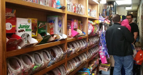A narrow candy shop aisle filled with colorful candy boxes and jars, with customers browsing and a child in an orange shirt.