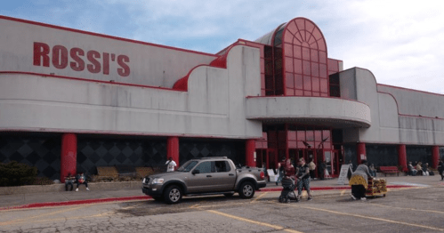 Exterior of Rossi's store with a red and gray facade, featuring shoppers and parked cars in the foreground.