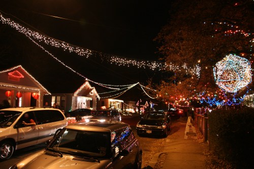 A festive street at night, adorned with colorful holiday lights on houses and trees, with parked cars lining the road.