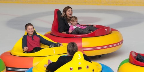 A woman and a girl smile while riding in a red bumper car on ice, with other colorful bumper cars nearby.