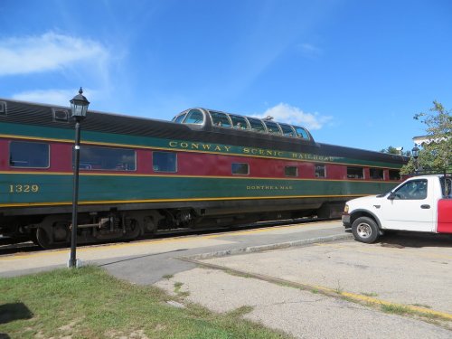 A vintage train car with a green and red exterior, labeled "Conway Scenic Railroad," parked beside a white truck.