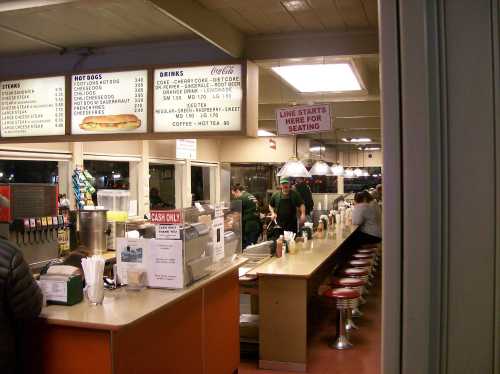 A diner interior with a counter, menu board, and patrons seated at red stools. A sign indicates where to line up for seating.