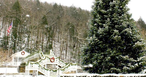 A snowy landscape featuring a large evergreen tree, a decorated gazebo, and a backdrop of wooded hills.
