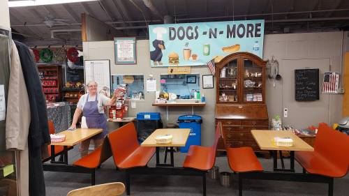 A cheerful woman stands in a cozy diner with a "Dogs -N- More" sign, surrounded by tables and a vintage decor.