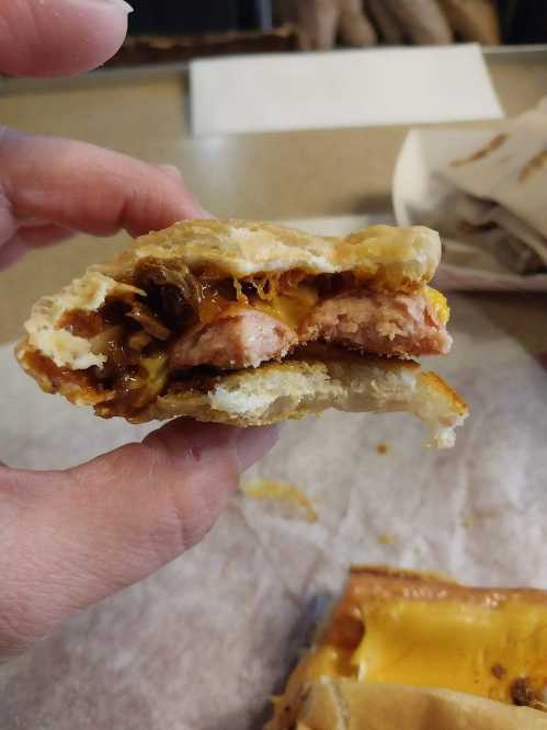 A hand holding a half-eaten sandwich with layers of meat, cheese, and sauce, resting on a paper surface.