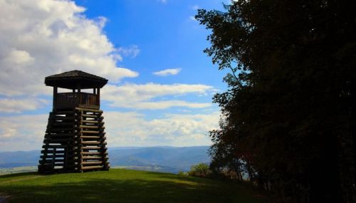 A wooden observation tower on a grassy hill, with a scenic view of mountains and a blue sky dotted with clouds.