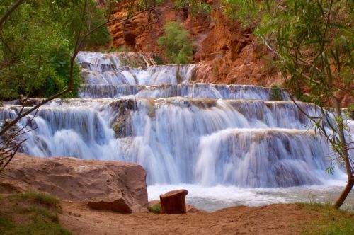 A serene waterfall cascades over rocky steps, surrounded by lush greenery and red rock formations.