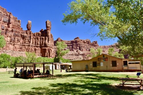 A scenic view of a grassy area with a building and picnic tables, surrounded by towering red rock formations and trees.