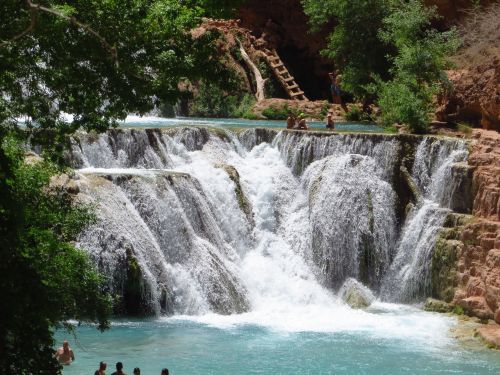 A scenic waterfall cascades into a turquoise pool, surrounded by lush greenery and people enjoying the water.