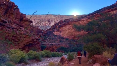 A scenic canyon with red rock formations and a bright sun rising above, with hikers walking along a gravel path.