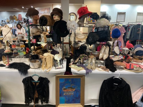 A vibrant display of vintage clothing, hats, accessories, and shoes on tables in a boutique setting.
