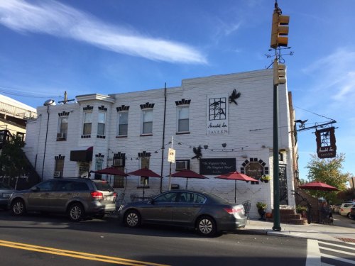 A white building with a sign reading "Tavern" and outdoor seating under red umbrellas on a sunny day.